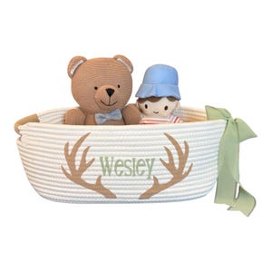 Peut inclure: Un panier en corde blanc avec le nom "Wesley" en vert et un motif de bois de cerf. Le panier contient un ours en peluche marron avec un n&oelig;ud papillon bleu et une poup&eacute;e portant un chapeau bleu et une chemise ray&eacute;e. Un ruban vert est nou&eacute; sur le c&ocirc;t&eacute;.