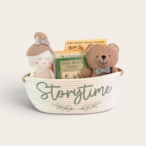 Puede incluir: Una cesta de cuerda blanca con la etiqueta "Storytime" contiene una mu&ntilde;eca, un oso de peluche y libros. La mu&ntilde;eca tiene un lazo y el oso lleva una pajarita. Los libros incluyen "How Do" y "Little Bear's Friend". La cesta tiene un dise&ntilde;o de hojas verdes.