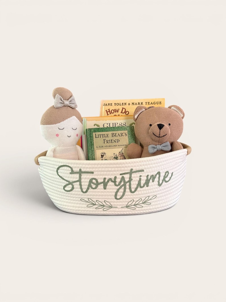 Puede incluir: Una cesta de cuerda blanca con la etiqueta "Storytime" contiene una mu&ntilde;eca, un oso de peluche y libros. La mu&ntilde;eca tiene un lazo y el oso lleva una pajarita. Los libros incluyen "How Do" y "Little Bear's Friend". La cesta tiene un dise&ntilde;o de hojas verdes.
