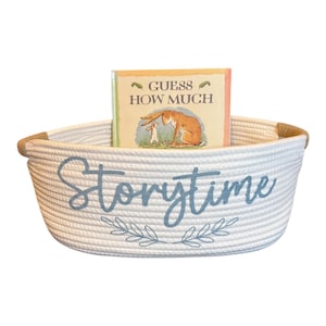 Puede incluir: Una cesta de cuerda blanca con la palabra "Storytime" en escritura azul y una ramita de hojas. Dentro de la cesta hay un libro infantil titulado "Adivina cu&aacute;nto te quiero". La cesta tiene un borde de cuerda marr&oacute;n.