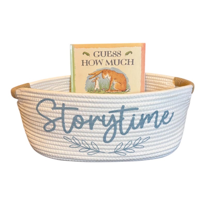 Puede incluir: Una cesta de cuerda blanca con la palabra "Storytime" en escritura azul y una ramita de hojas. Dentro de la cesta hay un libro infantil titulado "Adivina cu&aacute;nto te quiero". La cesta tiene un borde de cuerda marr&oacute;n.