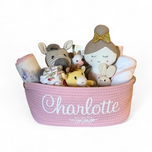 Peut inclure: Un panier rose tress&eacute; rempli d'articles pour b&eacute;b&eacute;s. Le panier contient une poup&eacute;e, un z&egrave;bre, une girafe, un lapin et une couverture. Le nom "Charlotte" est brod&eacute; sur le devant du panier en blanc.