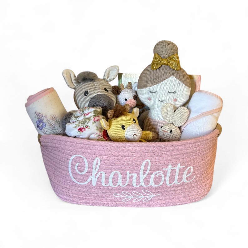Peut inclure: Un panier rose tress&eacute; rempli d'articles pour b&eacute;b&eacute;s. Le panier contient une poup&eacute;e, un z&egrave;bre, une girafe, un lapin et une couverture. Le nom "Charlotte" est brod&eacute; sur le devant du panier en blanc.