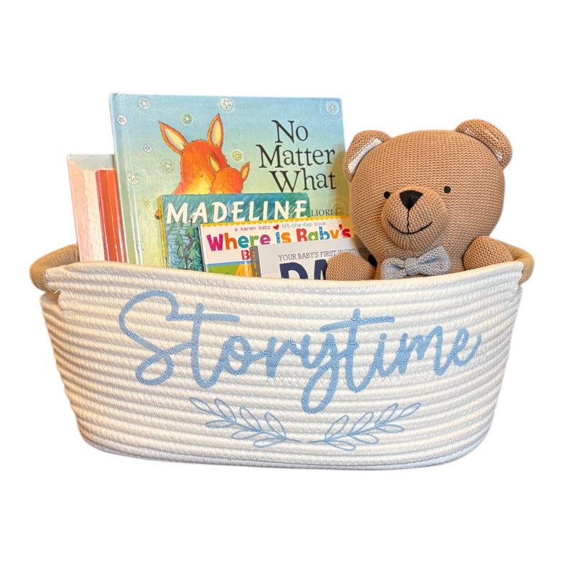 Puede incluir: Una cesta de cuerda blanca con la palabra "Storytime" en escritura azul claro, llena de libros infantiles y un oso de peluche marr&oacute;n claro. La cesta tiene un borde marr&oacute;n claro y un dise&ntilde;o floral azul.