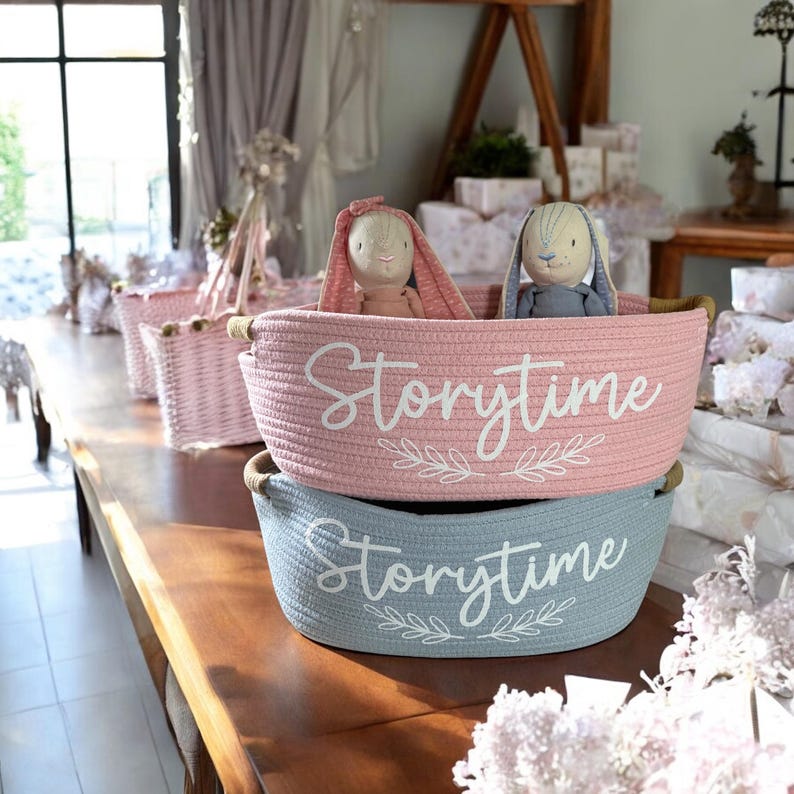 Puede incluir: Dos cestas de cuerda, una rosa y otra azul, con la palabra "Storytime" en escritura blanca. Cada cesta contiene un conejito de peluche. Se ven cestas rosas adicionales en el fondo.