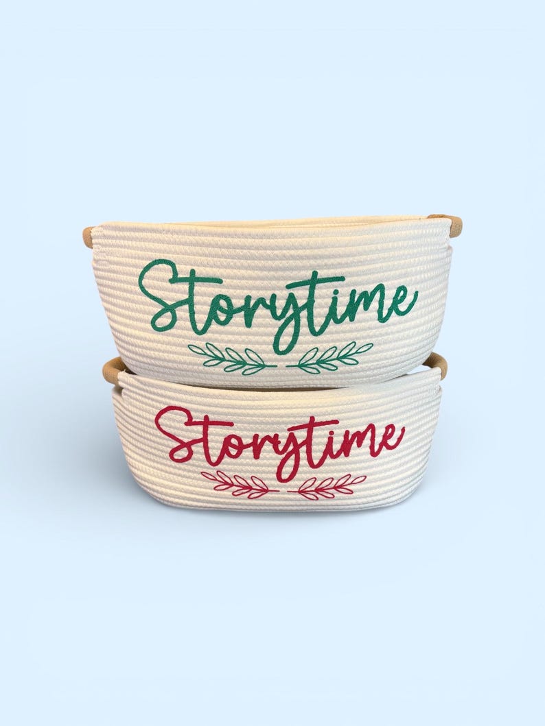 Puede incluir: Dos cestas de cuerda blanca apiladas. La cesta superior tiene la palabra "Storytime" en escritura verde con detalles de hojas verdes. La cesta inferior tiene "Storytime" en escritura roja con detalles de hojas rojas. Las cestas tienen asas de madera.