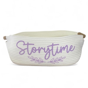 Puede incluir: Cesta de almacenamiento tejida blanca con la palabra "Storytime" en escritura p&uacute;rpura. La cesta tiene forma rectangular con esquinas redondeadas y asas de cuerda. Un dise&ntilde;o de hojas decorativas est&aacute; debajo de la palabra.