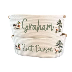 Peut inclure: Deux paniers en corde blanche avec des poign&eacute;es marron. Chaque panier porte un nom en &eacute;criture verte, ainsi qu'un canard et un cerf. Un panier dit "Graham" et l'autre "Rhett Dawson".