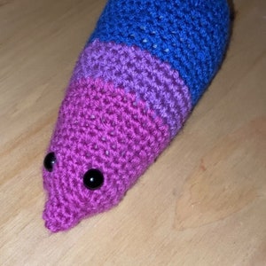 May include: A crocheted plush toy shaped like a worm with a pink, purple and blue colour scheme. The toy has two black eyes.