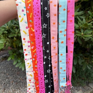 K&ouml;nnte beinhalten: Eine Sammlung von sieben Stoffb&auml;ndern mit verschiedenen bunten Mustern, darunter geometrische Formen, Sterne, Herzen und Monde in den Farben Wei&szlig;, Orange, Pink, Schwarz, Hellrosa und Hellblau, jeweils mit einem silbernen Metallverschluss versehen.
