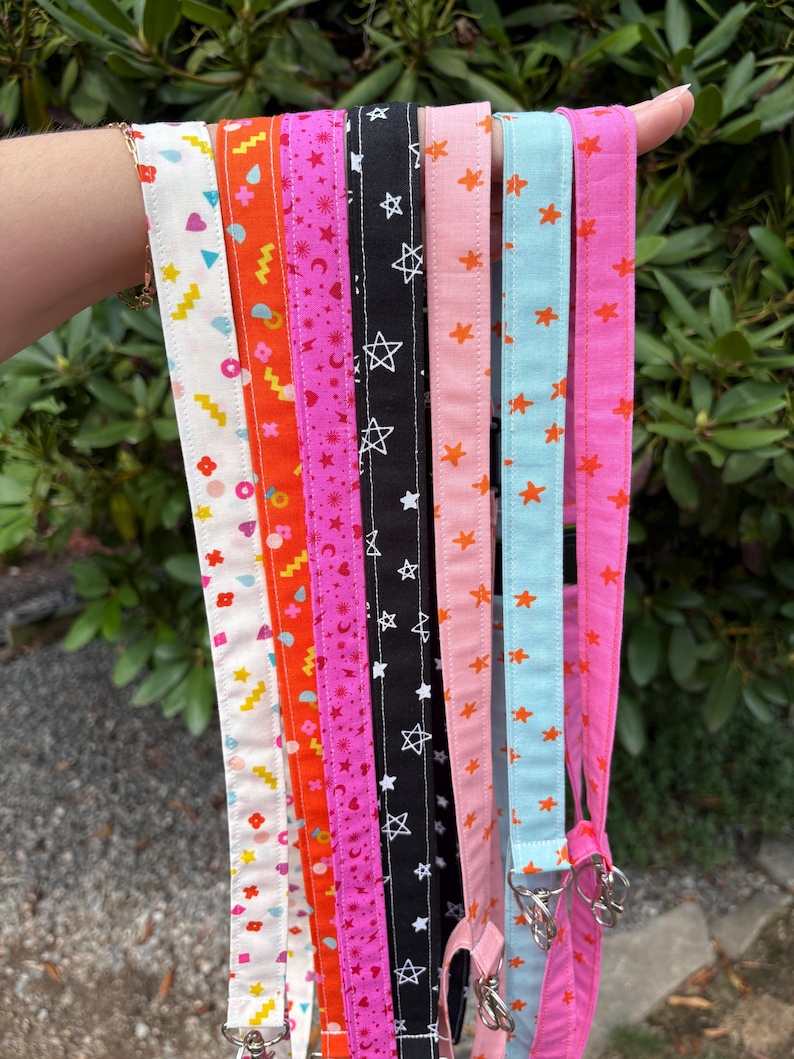 K&ouml;nnte beinhalten: Eine Sammlung von sieben Stoffb&auml;ndern mit verschiedenen bunten Mustern, darunter geometrische Formen, Sterne, Herzen und Monde in den Farben Wei&szlig;, Orange, Pink, Schwarz, Hellrosa und Hellblau, jeweils mit einem silbernen Metallverschluss versehen.
