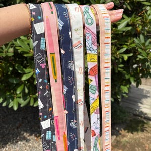 K&ouml;nnte beinhalten: Eine Sammlung von sechs Stoffb&auml;ndern mit verschiedenen Mustern von Schul- und B&uuml;robedarf. Die Designs zeigen Illustrationen von Stiften, Scheren, Linealen, B&uuml;roklammern und Umschl&auml;gen in Farben wie Schwarz, Pink, Marineblau, Wei&szlig; und Gelb.