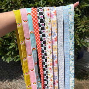 K&ouml;nnte beinhalten: Eine Sammlung von zehn Stoff-Lanyards mit verschiedenen Mustern, darunter Gelb, Rosa mit Regenb&ouml;gen, Hellblau mit Regenb&ouml;gen, orangefarbenes Karomuster, schwarz-wei&szlig;es Karomuster mit Rosen, Blumenmuster und blaue G&auml;nsebl&uuml;mchen-Designs mit Metallclips.
