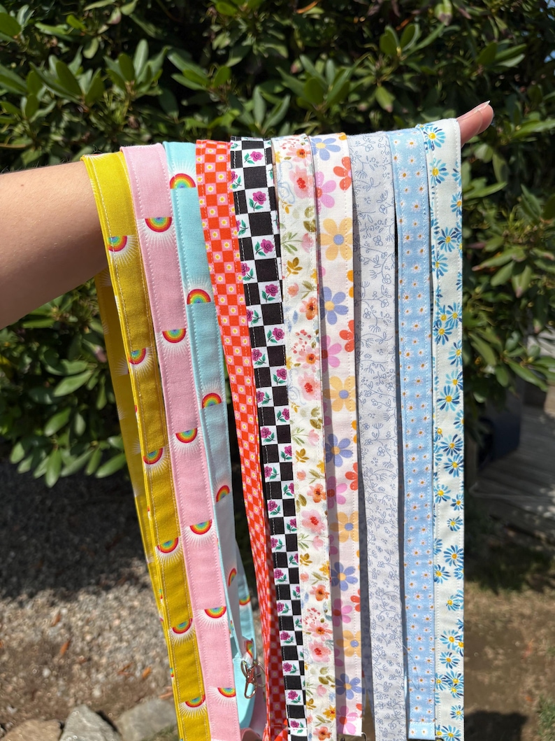 K&ouml;nnte beinhalten: Eine Sammlung von zehn Stoff-Lanyards mit verschiedenen Mustern, darunter Gelb, Rosa mit Regenb&ouml;gen, Hellblau mit Regenb&ouml;gen, orangefarbenes Karomuster, schwarz-wei&szlig;es Karomuster mit Rosen, Blumenmuster und blaue G&auml;nsebl&uuml;mchen-Designs mit Metallclips.