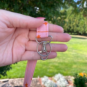 K&ouml;nnte beinhalten: Ein rosa Stoff-Lanyard mit einem sich wiederholenden Muster aus kleinen orangefarbenen Sternen. Das Lanyard ist an einem silberfarbenen Metall-Karabinerhaken und einem runden Schl&uuml;sselring befestigt, die als sichere Halterung f&uuml;r Schl&uuml;ssel dienen.