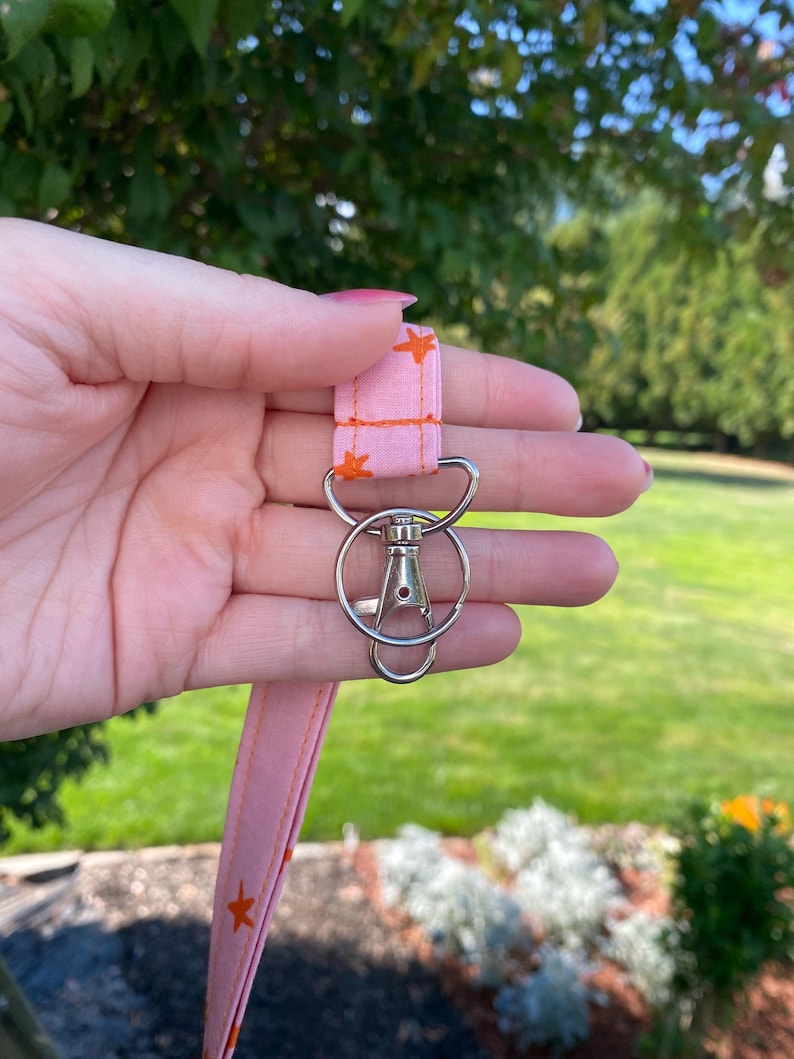 K&ouml;nnte beinhalten: Ein rosa Stoff-Lanyard mit einem sich wiederholenden Muster aus kleinen orangefarbenen Sternen. Das Lanyard ist an einem silberfarbenen Metall-Karabinerhaken und einem runden Schl&uuml;sselring befestigt, die als sichere Halterung f&uuml;r Schl&uuml;ssel dienen.