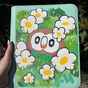 May include: A light mint green binder featuring a brown and white owl character with a small orange beak, surrounded by white flowers with yellow centers and green grass. The design includes small white star accents and a green logo in the corner.