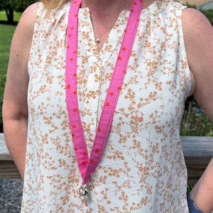 K&ouml;nnte beinhalten: Ein leuchtend pinkfarbenes Stoff-Lanyard mit einem sich wiederholenden Muster aus kleinen orangefarbenen Sternen. Das Lanyard besteht aus einem flachen Textilband und verf&uuml;gt &uuml;ber einen silberfarbenen Metall-Drehverschluss zum sicheren Befestigen.