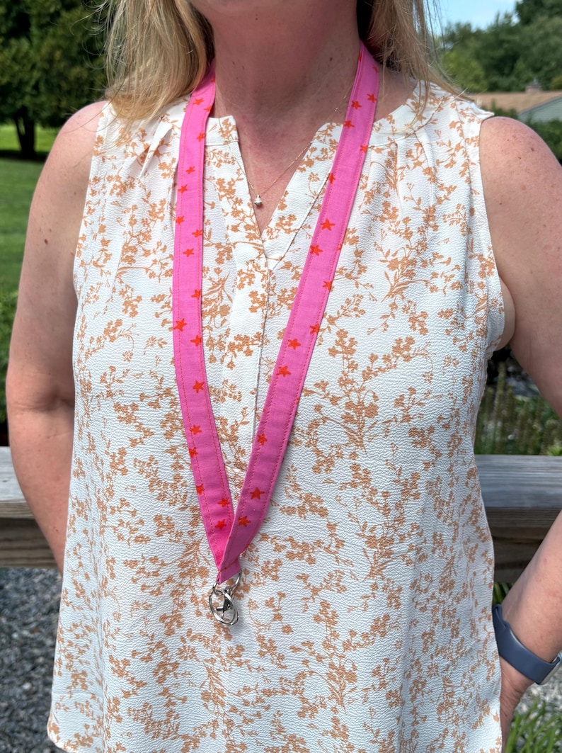 K&ouml;nnte beinhalten: Ein leuchtend pinkfarbenes Stoff-Lanyard mit einem sich wiederholenden Muster aus kleinen orangefarbenen Sternen. Das Lanyard besteht aus einem flachen Textilband und verf&uuml;gt &uuml;ber einen silberfarbenen Metall-Drehverschluss zum sicheren Befestigen.