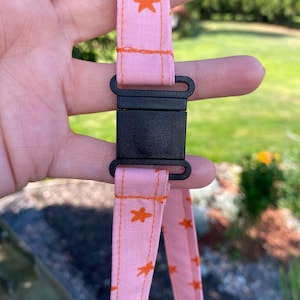 K&ouml;nnte beinhalten: Ein hellrosa Stoffband mit einem sich wiederholenden Muster aus kleinen orangefarbenen Sternen. Das Band ist mit einer schwarzen Kunststoff-Steckschnalle gesichert, wobei orangefarbene N&auml;hte entlang der Stoffkanten sichtbar sind.