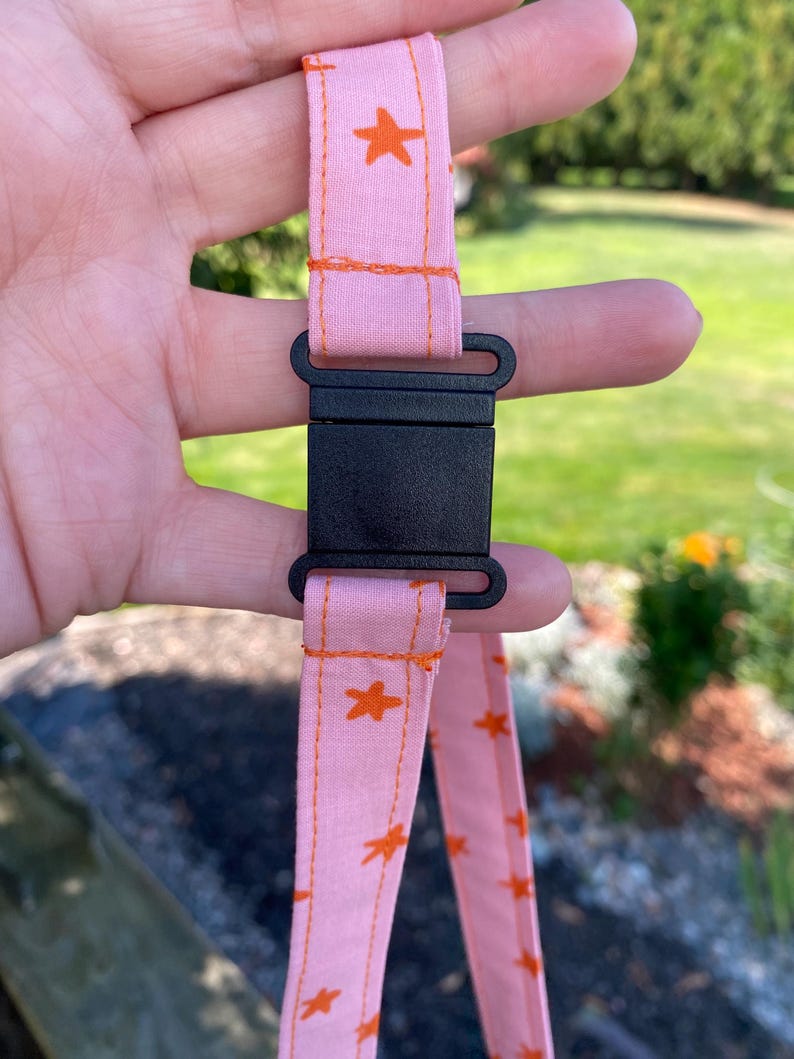 K&ouml;nnte beinhalten: Ein hellrosa Stoffband mit einem sich wiederholenden Muster aus kleinen orangefarbenen Sternen. Das Band ist mit einer schwarzen Kunststoff-Steckschnalle gesichert, wobei orangefarbene N&auml;hte entlang der Stoffkanten sichtbar sind.