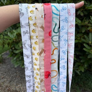K&ouml;nnte beinhalten: Eine Sammlung von sieben Stoffb&auml;ndern mit verschiedenen Mustern, darunter schwarze Schmetterlinge, gelbe Quietscheentchen, braune Hunde, rote Erdbeeren, t&uuml;rkisfarbene Kringel, blaue Schleifen und rosa Schleifen, die vertikal angeordnet sind.