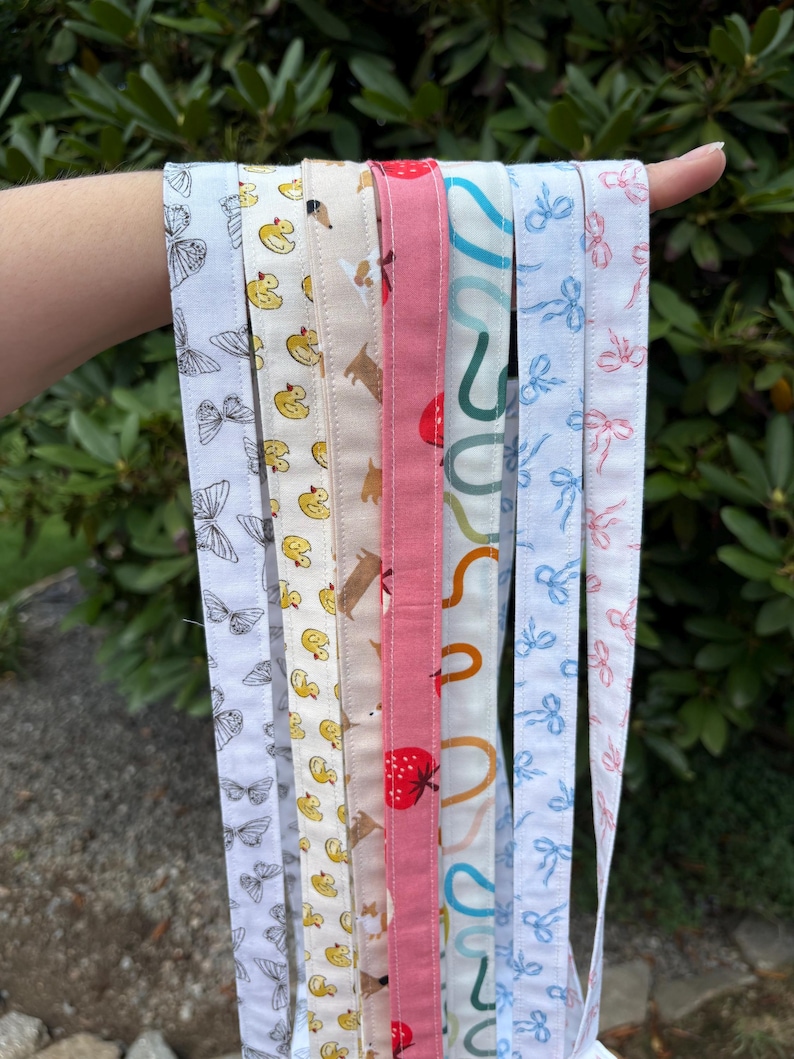 K&ouml;nnte beinhalten: Eine Sammlung von sieben Stoffb&auml;ndern mit verschiedenen Mustern, darunter schwarze Schmetterlinge, gelbe Quietscheentchen, braune Hunde, rote Erdbeeren, t&uuml;rkisfarbene Kringel, blaue Schleifen und rosa Schleifen, die vertikal angeordnet sind.