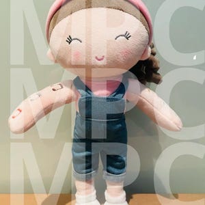Puede incluir: Muñeca de peluche con una cara sonriente, que lleva una diadema rosa, un peto vaquero y zapatos blancos. La muñeca tiene el pelo castaño y una camisa rosa. Un tatuaje de nota musical es visible en el brazo de la muñeca.