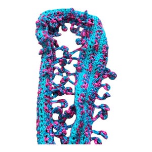 Puede incluir: Bufanda de ganchillo hecha a mano en una vibrante combinación de colores turquesa y fucsia. La bufanda presenta un diseño abierto y calado con bordes decorativos y rizados. El hilo parece ser un material suave y texturizado, perfecto para añadir color.