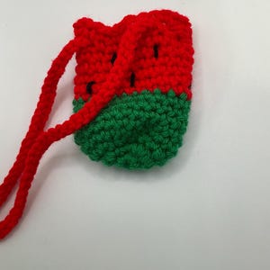 May include: A small, handmade crochet pouch in the shape of a watermelon. The top half is red with black seed details, and the bottom half is green. A red cord is attached for carrying.