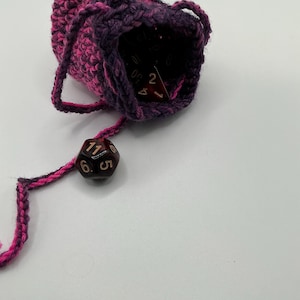 May include: A handmade crochet dice bag in shades of pink and purple. The bag is open, revealing several dice inside. A single die is attached to the bag by a crocheted string. The dice are black with red numbers. The bag is designed to hold dice for tabletop games.