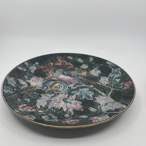 May include: Decorative plate with a dark green background and a detailed floral and bird design. The plate features a gold rim and a colorful illustration of a bird and flowers in shades of pink, blue, and white.