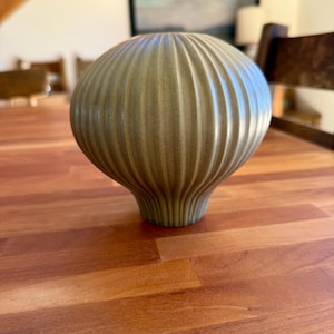 May include: A decorative, olive-green ceramic vase with a ribbed, bulbous body and a flared base. The vase is approximately 8 inches tall and sits on a wooden surface. The background includes a painting and furniture.