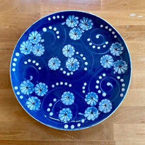 May include: A blue ceramic plate with a white floral design. The plate features white flowers with brown centers and white swirls on a dark blue background.