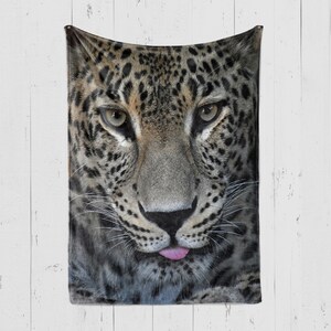 May include: Close-up of a leopard's face with black spots on a tan background. The leopard is looking directly at the camera with its mouth slightly open, revealing a pink tongue.