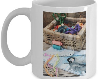 May include: White ceramic mug with a colorful image of a wicker basket filled with embroidery floss, scissors, and spools of thread. A cross-stitch pattern is visible on a wooden surface.