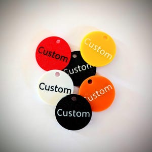 May include: A collection of six circular acrylic tags in red, yellow, black, white, and orange. Each tag features a small hole at the top and the word Custom printed in a contrasting color. The tags are arranged in a cluster on a plain surface.