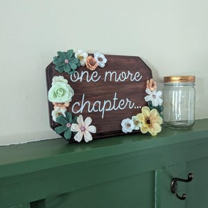 May include: A wooden sign with the text "one more chapter..." surrounded by artificial flowers in shades of green, white, and yellow. The sign is sitting on a green shelf.