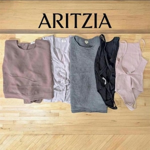 May include: A collection of women's clothing items, including a mauve-colored sweatshirt, a light gray blouse, a gray ribbed tank top, a black bodysuit, and a pink ribbed bodysuit. The brand name "ARITZIA" is displayed in large, black capital letters.