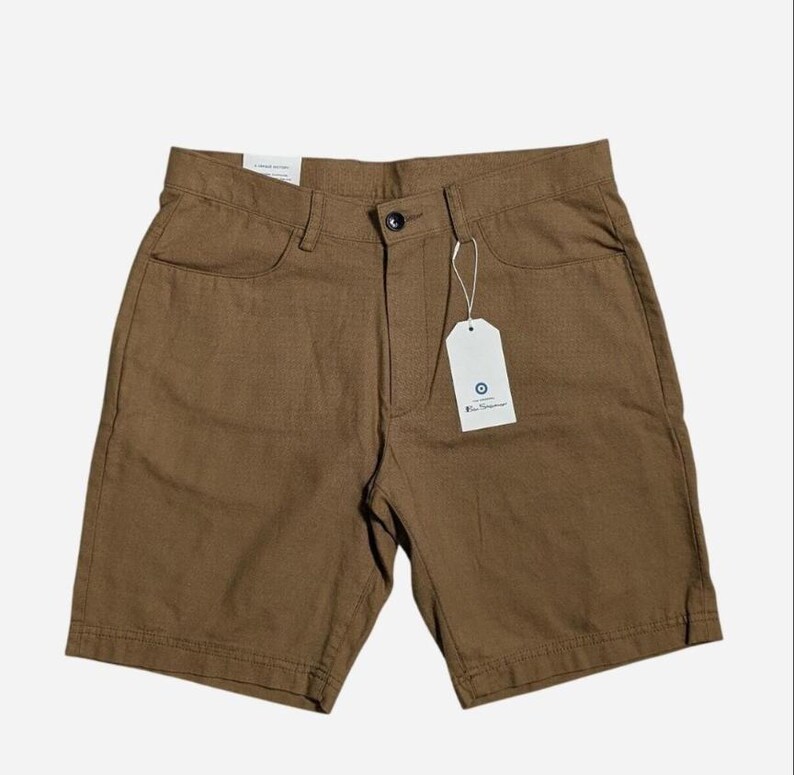 May include: Brown shorts with a button closure, front pockets, and a tag. The shorts are made of a textured fabric and have a classic design. The tag has a blue circle logo. These shorts are suitable for casual wear.