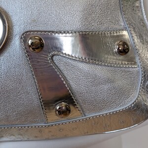 Puede incluir: Bolso plateado con acabado de cuero texturizado. El bolso presenta detalles plateados pulidos, incluido un panel decorativo con tres tachuelas redondas plateadas. El dise&ntilde;o del bolso incluye un borde cosido.