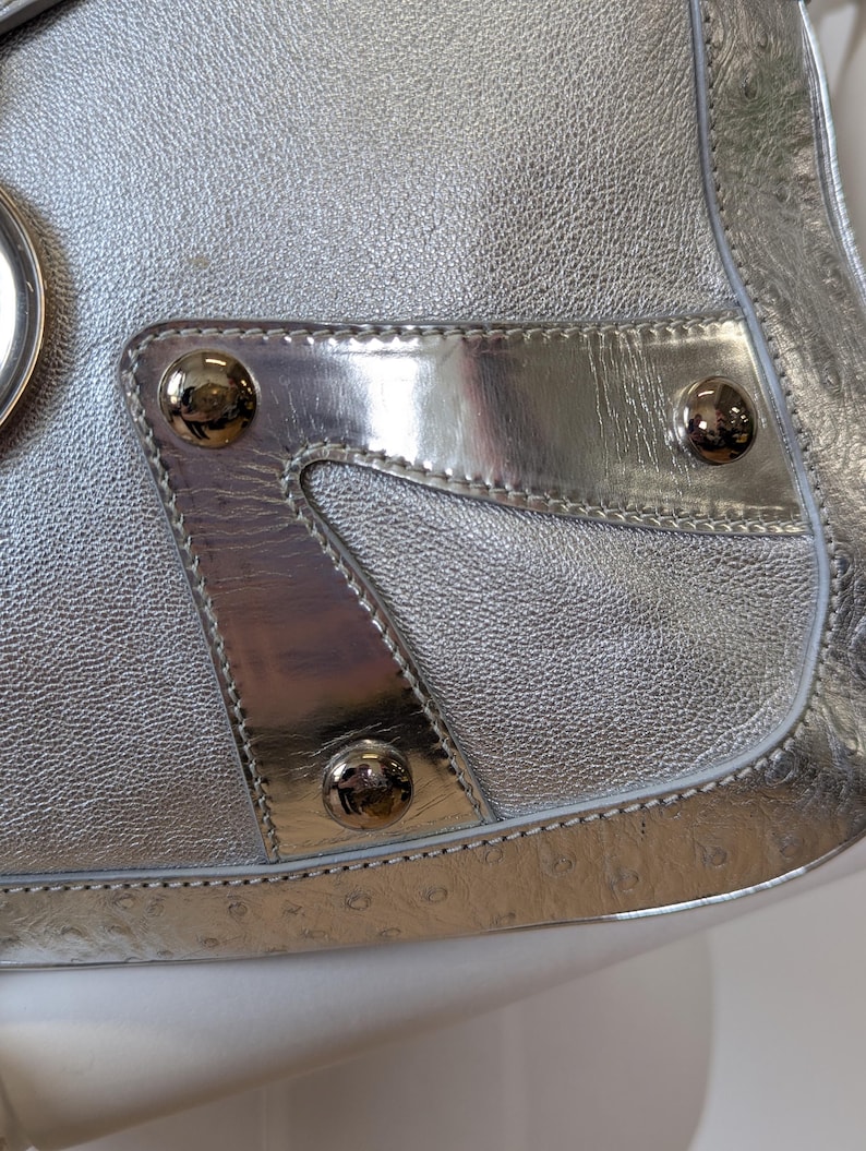Puede incluir: Bolso plateado con acabado de cuero texturizado. El bolso presenta detalles plateados pulidos, incluido un panel decorativo con tres tachuelas redondas plateadas. El dise&ntilde;o del bolso incluye un borde cosido.