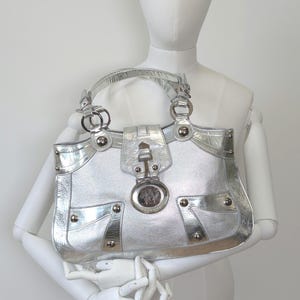 Puede incluir: Bolso de mano plateado con acabado brillante. El bolso presenta detalles plateados, incluyendo un cierre decorativo, remaches y un emblema con logotipo. Tiene un asa superior y un dise&ntilde;o estructurado.