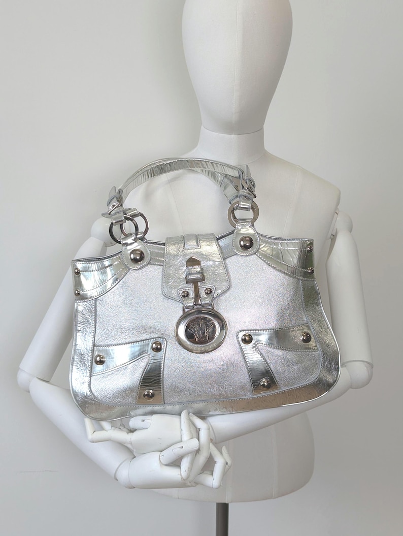 Puede incluir: Bolso de mano plateado con acabado brillante. El bolso presenta detalles plateados, incluyendo un cierre decorativo, remaches y un emblema con logotipo. Tiene un asa superior y un dise&ntilde;o estructurado.