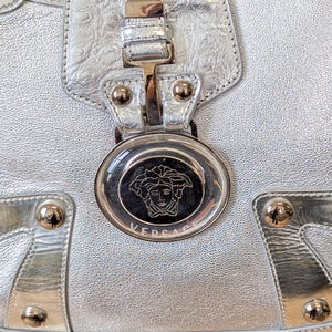Puede incluir: Primer plano de un bolso de mano de cuero plateado con una superficie texturizada. El bolso presenta herrajes dorados, incluyendo tachuelas y un cierre. Es visible un emblema circular con la marca "VERSACE".