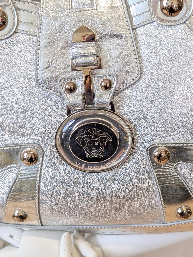 Puede incluir: Primer plano de un bolso de mano de cuero plateado con una superficie texturizada. El bolso presenta herrajes dorados, incluyendo tachuelas y un cierre. Es visible un emblema circular con la marca "VERSACE".