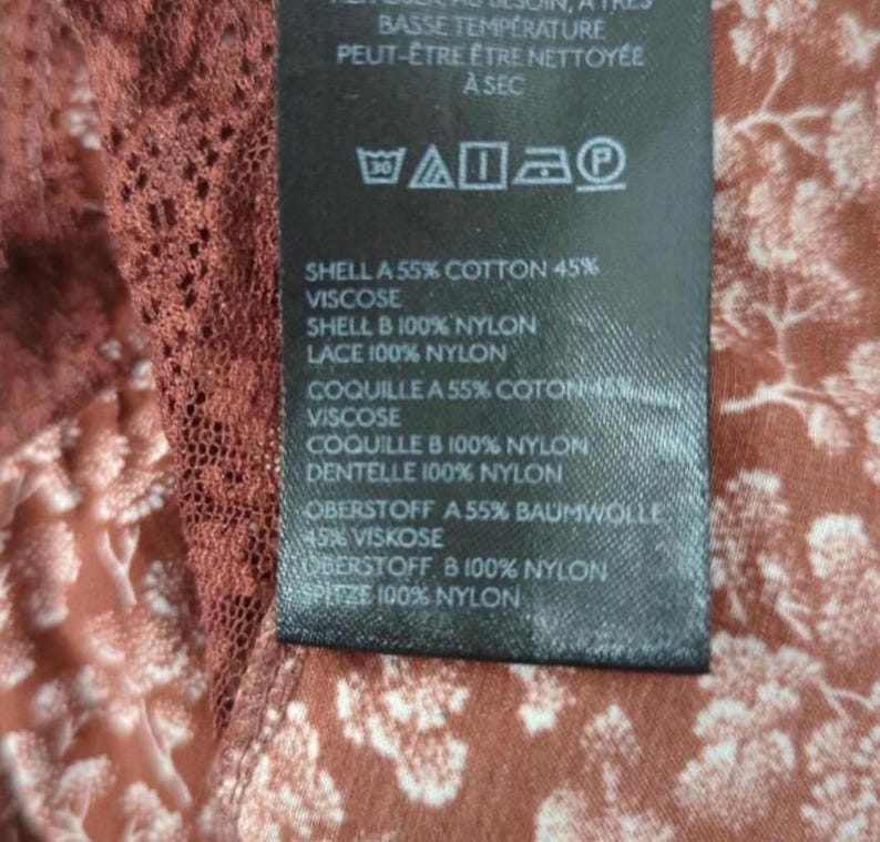 May include: A close-up of a clothing label with text in English and French. The label details fabric content, such as cotton, viscose, and nylon. The background features a floral pattern in brown and white.