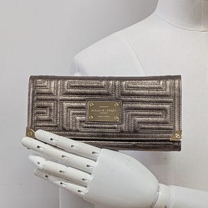 Może przedstawiać: Metaliczny, brązowy portfel z geometrycznym wzorem i złotymi akcentami. Prostokątny portfel posiada zapięcie na klapę i małą złotą plakietkę z napisem "GIANNI VERSACE COUTURE".