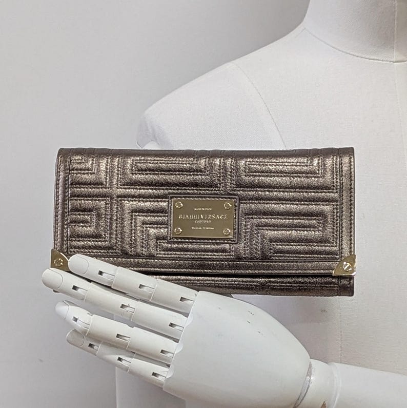 May include: A metallic bronze wallet with a geometric pattern and gold-toned accents. The wallet features a rectangular shape with a flap closure and a small gold plaque with the text "GIANNI VERSACE COUTURE".