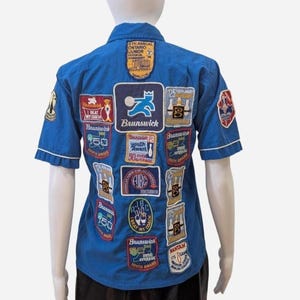 May include: A blue short-sleeved shirt with white trim, covered in embroidered patches. The patches feature bowling-related imagery, the Brunswick logo, and text including "Youth Award" and "Beat My Coach".